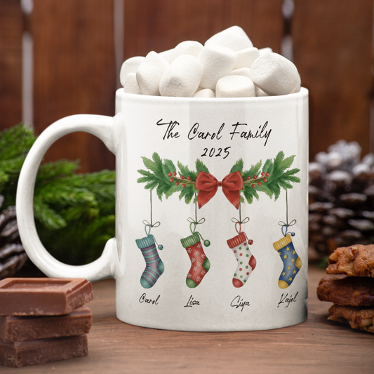 White mug with Christmas-themed design featuring stockings and a bow, filled with hot chocolate and marshmallows, on a wooden surface.