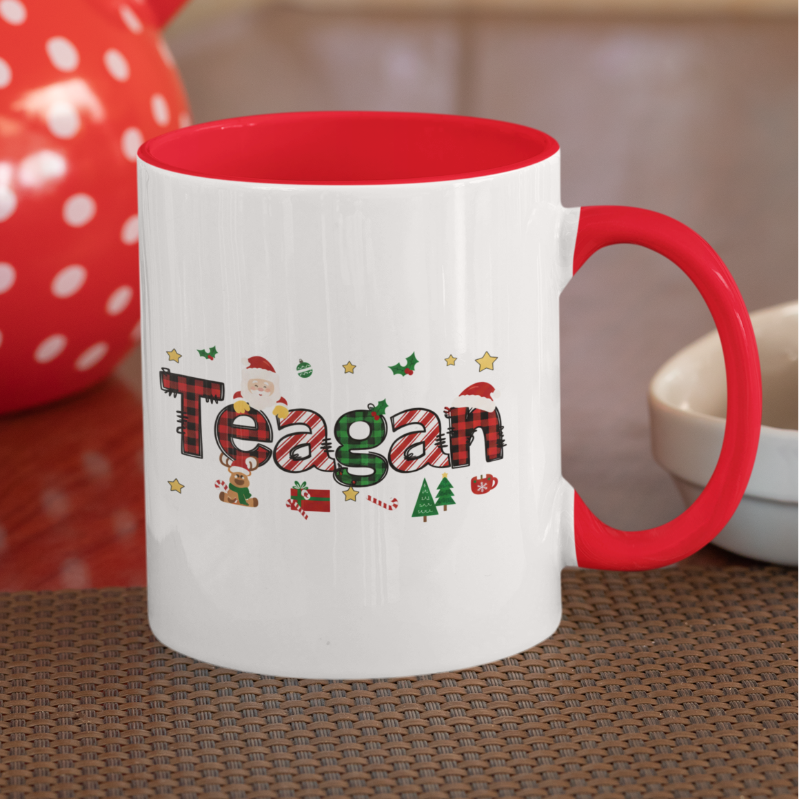 White mug with red interior and handle, featuring the name 'Teagan' in colorful letters, placed on a wooden surface with a red polka dot teapot in the background.